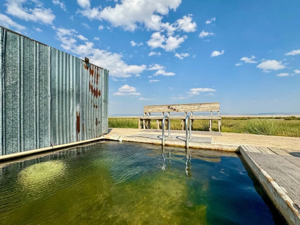 A serene outdoor saltwater pool surrounded by tall corrugated walls under a bright blue sky with scattered clouds. The clear greenish-blue water reflects the sky and surrounding landscape.