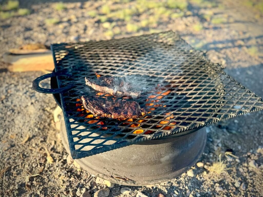 A close-up view of a portable metal grill over a campfire with chopped wood nearby.
