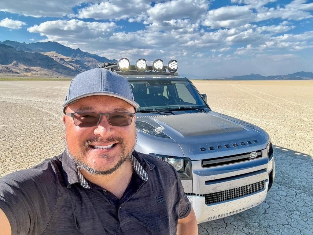 A smiling man poses with a top-mounted light-equipped Land Rover on cracked desert ground under a open sky, with distant mountains and scattered clouds.