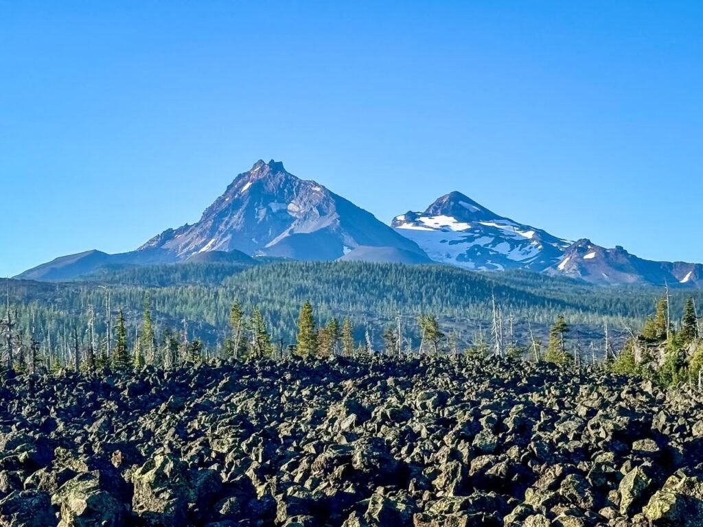 A rugged volcanic landscape features dark lava rocks in the foreground and snow-capped mountains under a clear blue sky.