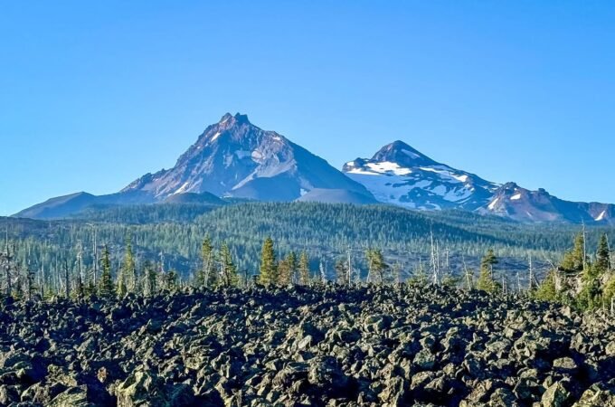 A rugged volcanic landscape features dark lava rocks in the foreground and snow-capped mountains under a clear blue sky.