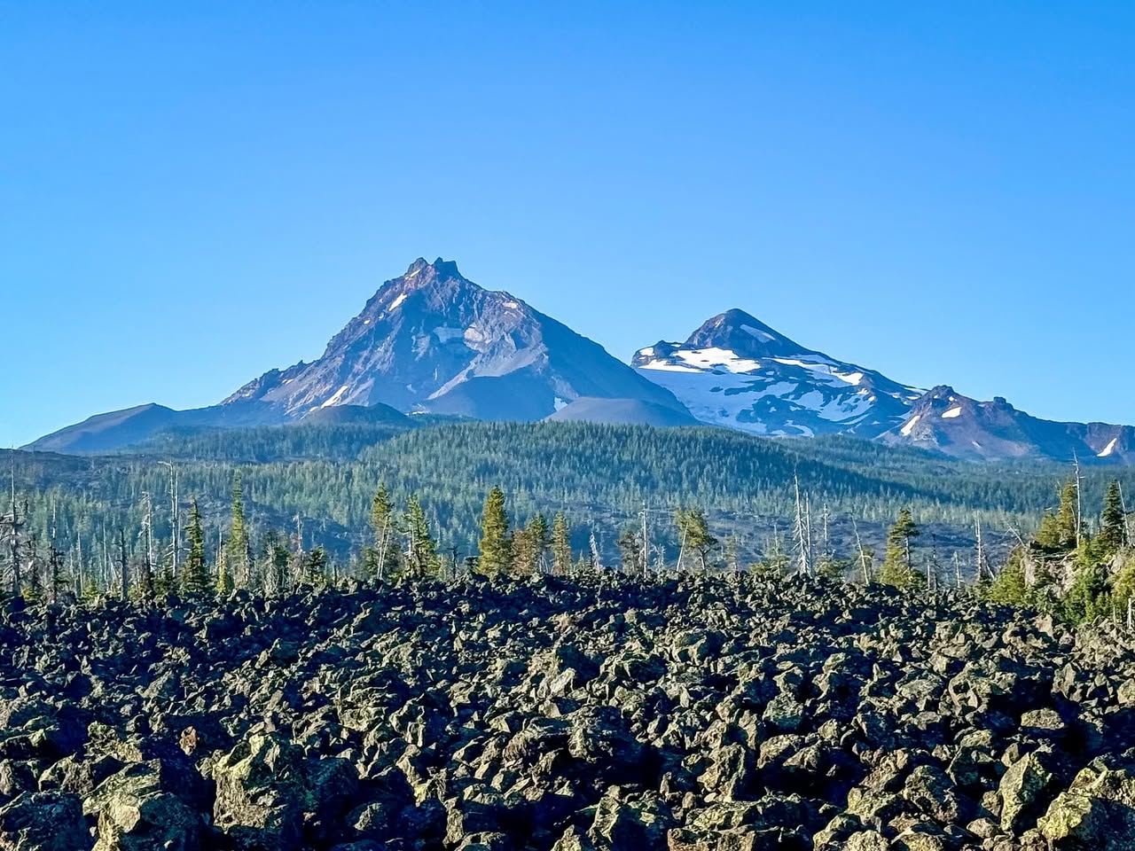 A rugged volcanic landscape features dark lava rocks in the foreground and snow-capped mountains under a clear blue sky.