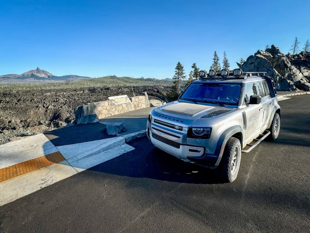 A silver SUV parked at a scenic overlook in a highlands landscape with a mountain in the background. Blue skies and a rugged valley fill the scene.