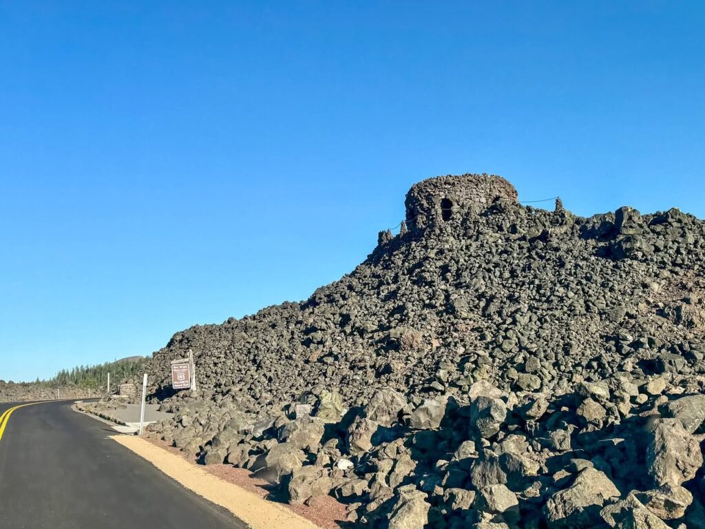 A rugged volcanic rock formation with a distinctive cave-like opening sits beside a paved road under a bright blue sky. The scene showcases natural textures and arid landscape features.