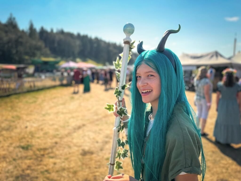 A person with teal hair and horns holds a vine-covered staff at an outdoor festival with tents in the background.