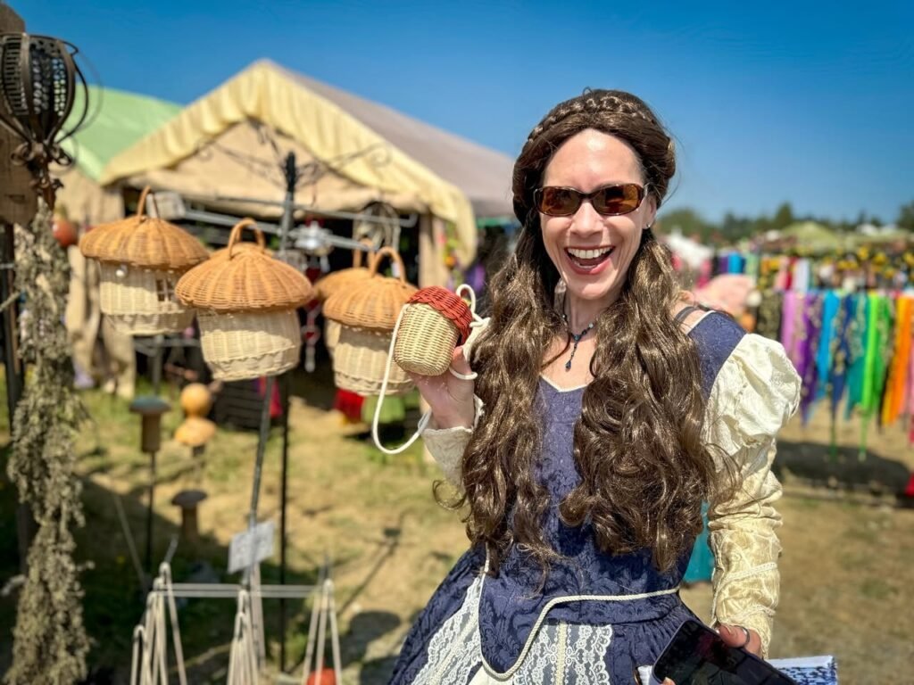 A smiling visitor holds a woven basket at an outdoor farm market. Bright colorful flags hang in the background under a sunny sky.