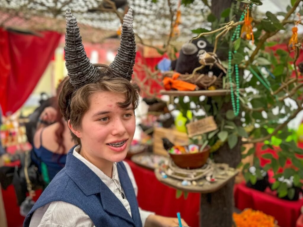 A person wearing a blue vest and large, gray, twisted ram horns stands near a decorated tree. The scene is set within a crowded street fair or outdoor market.