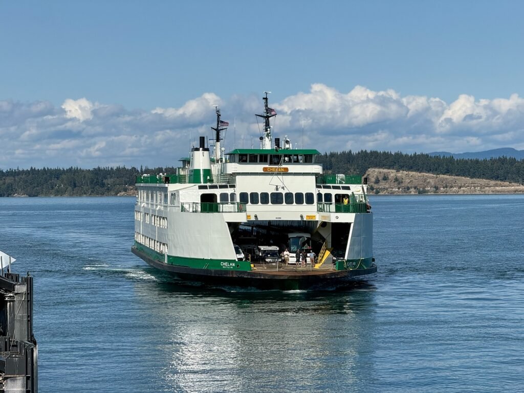 A green and white Washington State Ferry crosses calm, blue water with forested shores in the background.