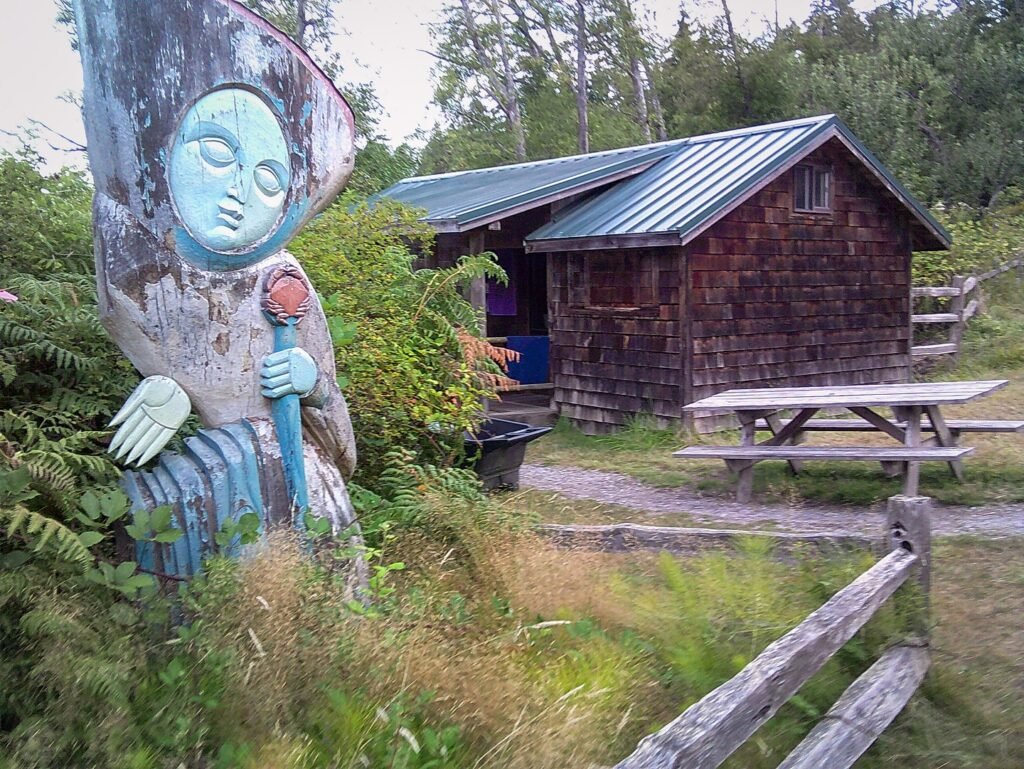 A weathered wooden cabin with a metal roof sits alongside a whimsical face sculpture and a rustic fence, surrounded by lush greenery.