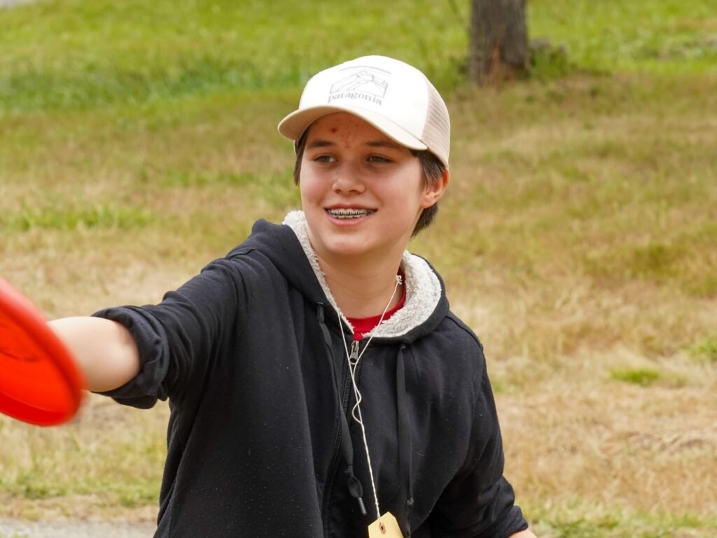 A person smiles while extending an arm holding a bright red frisbee. This shows a sunny day outdoors on a grassy field.