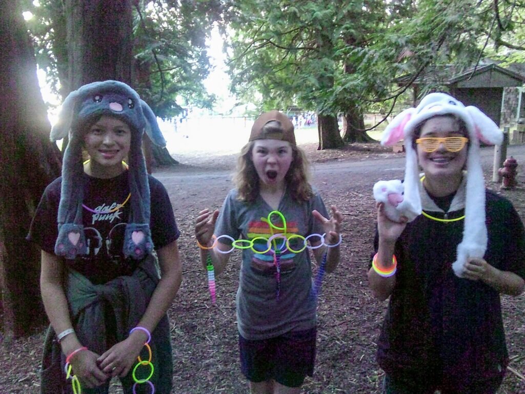 A group of people wearing animal hats and glow sticks in a forested area at a festival. One person smiles in a blue animal hat.