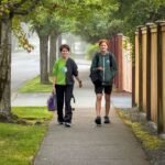 Two people walking on a sidewalk under a canopy of large trees. The scene is misty and green, with a wooden fence running alongside the path.