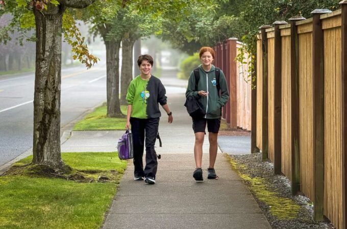 Two people walking on a sidewalk under a canopy of large trees. The scene is misty and green, with a wooden fence running alongside the path.