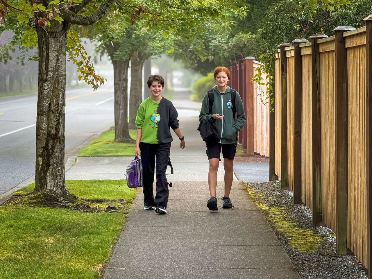 Two people walking on a sidewalk under a canopy of large trees. The scene is misty and green, with a wooden fence running alongside the path.