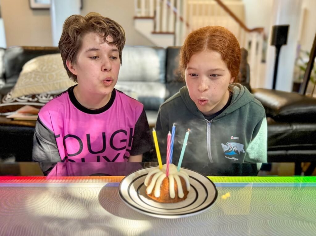 Two young people blowing out colorful candles on a small birthday cake in a home setting.