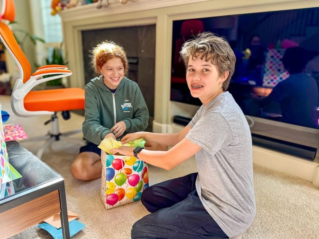 Two young boys share the joy of opening a colorful present on the carpet. The scene includes a fireplace, orange chair, and a television in the background.