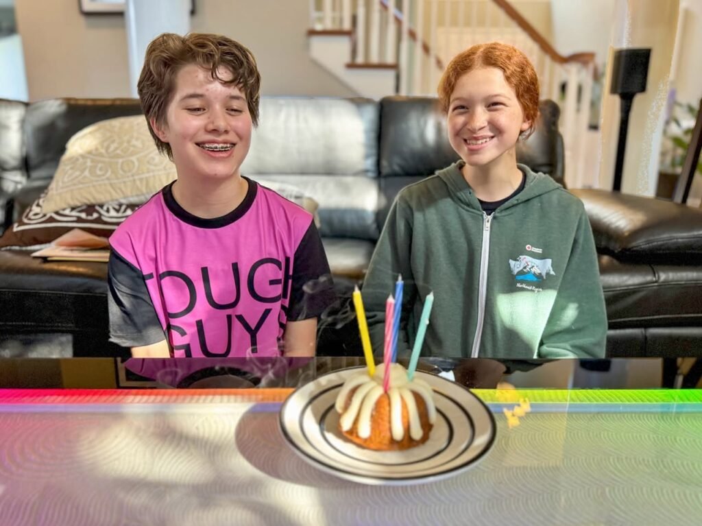 Two children smile at a birthday sundae with lit candles in a living room.