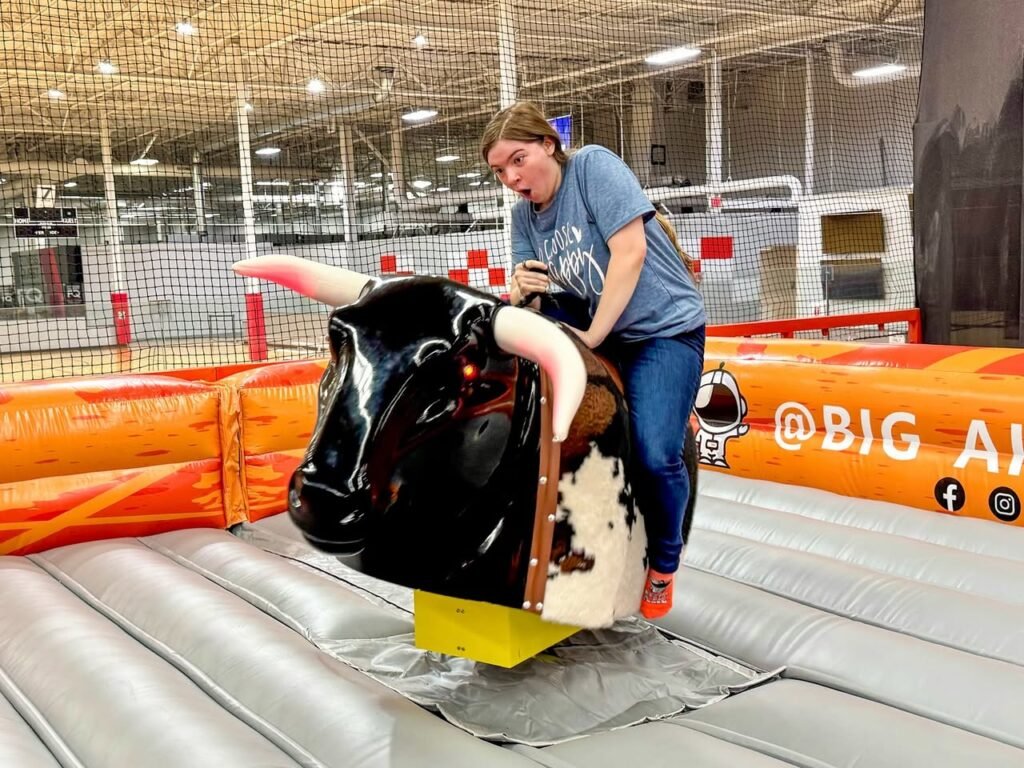 An excited person is riding a mechanical bull attraction at an indoor fair with an inflatable barrier.