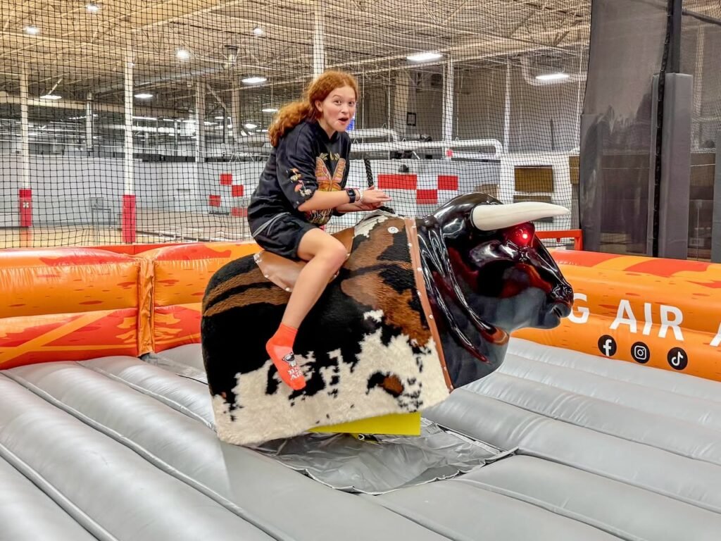 A person smiles while riding a mechanical bull at an indoor trampoline park surrounded by safety netting.