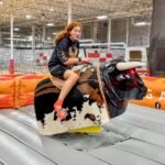 VR Birthday A person smiles while riding a mechanical bull at an indoor trampoline park surrounded by safety netting.