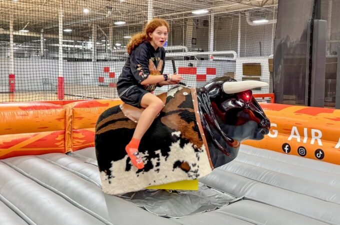 A person smiles while riding a mechanical bull at an indoor trampoline park surrounded by safety netting.