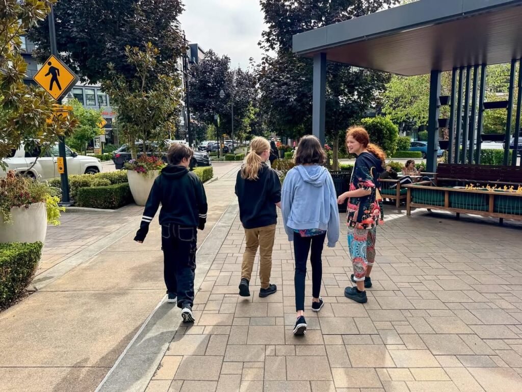 Four people walking away on a paved sidewalk near a bus stop with tall trees in the background.