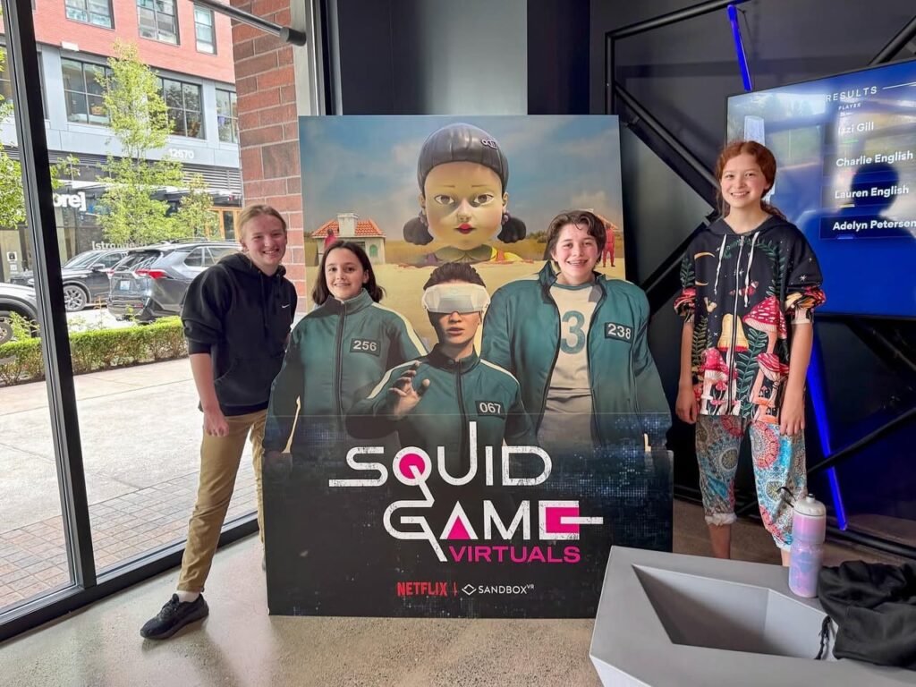 Individuals standing on a virtual reality platform with a Squid Game Virtals sign in front of large storefront windows.