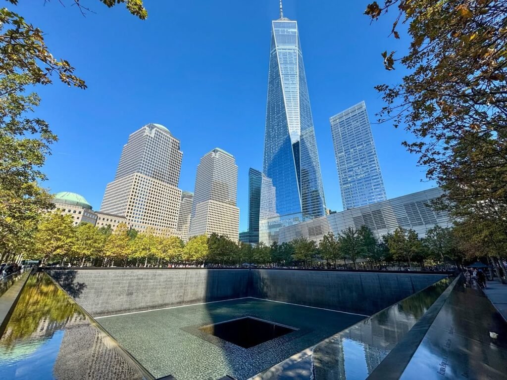 The square Memorial Pool at the National September 11 Memorial beneath the falling water. In the background, One World Trade Center stands tall against the clear blue sky.