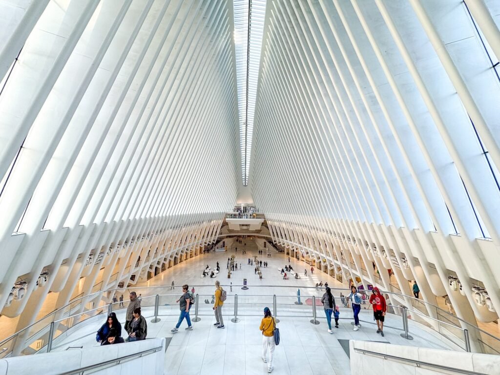 Visitors walk the terminal beneath the white, web-like structure of the Oculus. A distinctive spire is visible under the arched ceiling, set against bright, natural light.