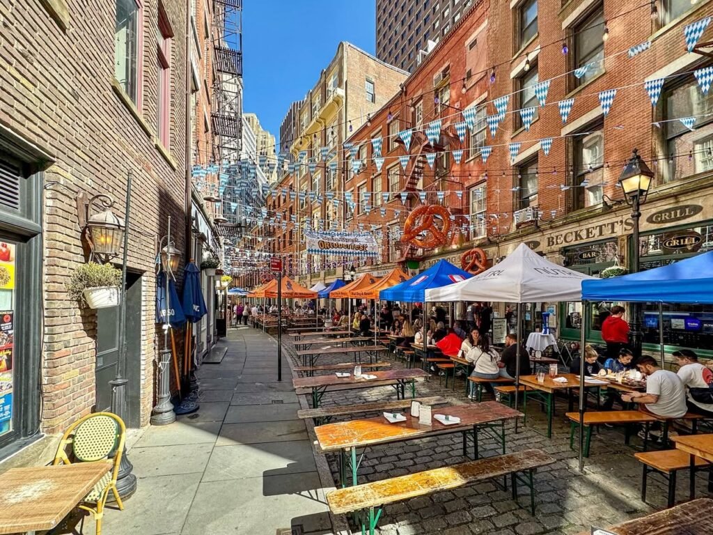 A bustling outdoor market with colorful tents, brick buildings, and people dining at wooden tables. Vibrant urban atmosphere with festive decorations.