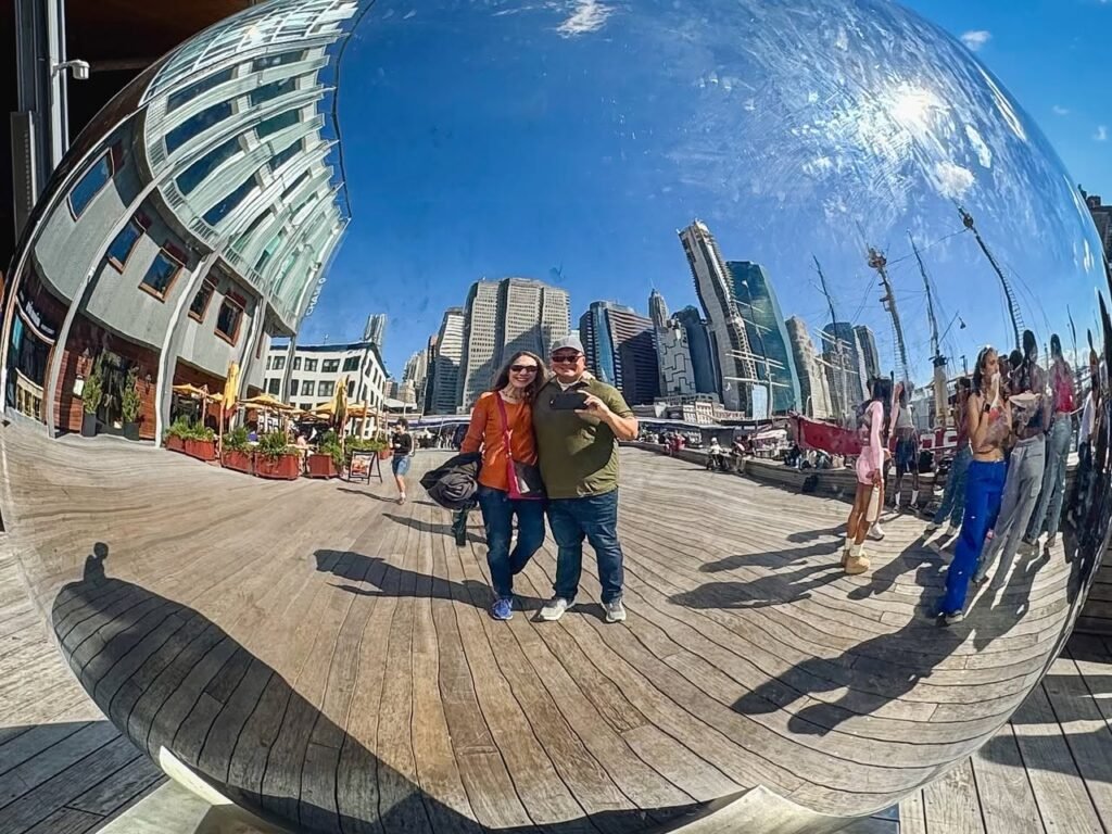 A couple poses with a selfie inside a giant silver sphere on the Seattle waterfront. The distorted mirror reflects the skyline and clear blue sky around them.