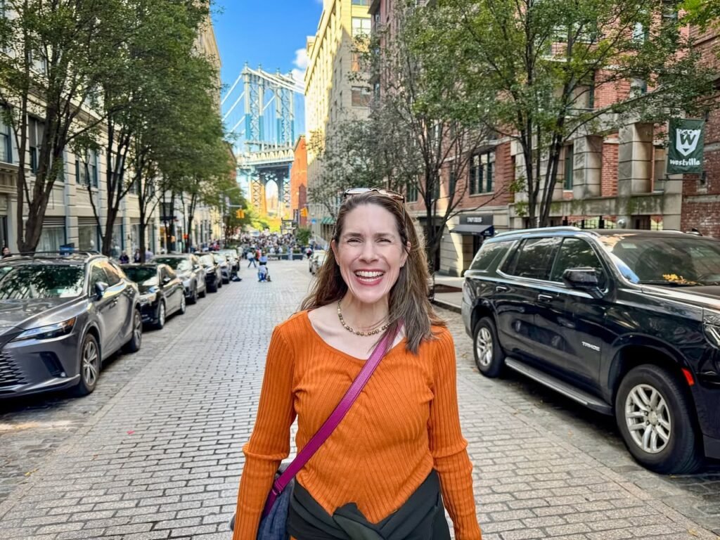 A woman in an orange top stands on a cobblestone road in San Francisco with the Richmond Bridge in the background. The photo captures a sunny urban scene with parked cars and brick buildings.