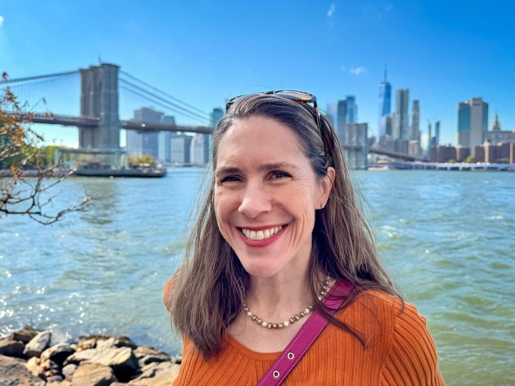 A smiling visitor poses at Brooklyn Bridge Park with the iconic bridge and NYC skyline in the background.