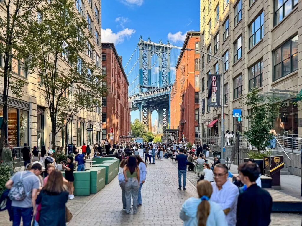 A bustling pedestrian street in DUMBO, Brooklyn, leading to a stunning view of the Manhattan Bridge. Multi-story brick and stone buildings line the street under a clear blue sky.