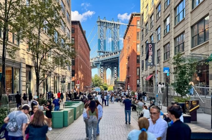 A bustling pedestrian street in DUMBO, Brooklyn, leading to a stunning view of the Manhattan Bridge. Multi-story brick and stone buildings line the street under a clear blue sky.