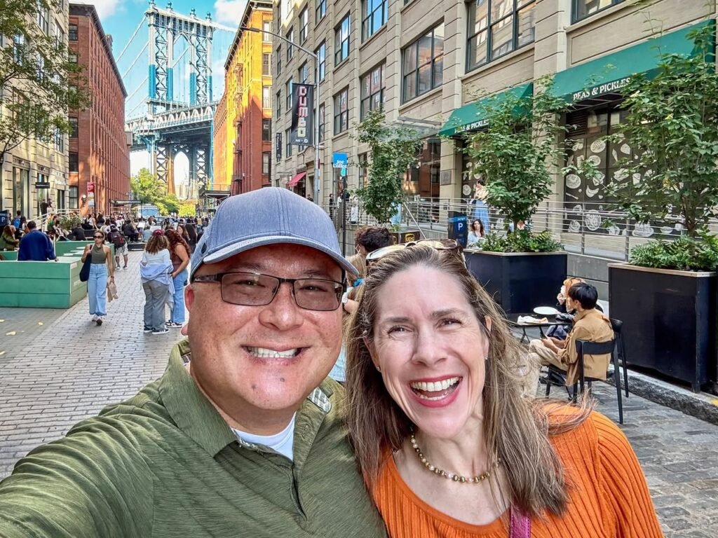 A couple taking a smiling selfie on a New York street with the Brooklyn Bridge in the background.