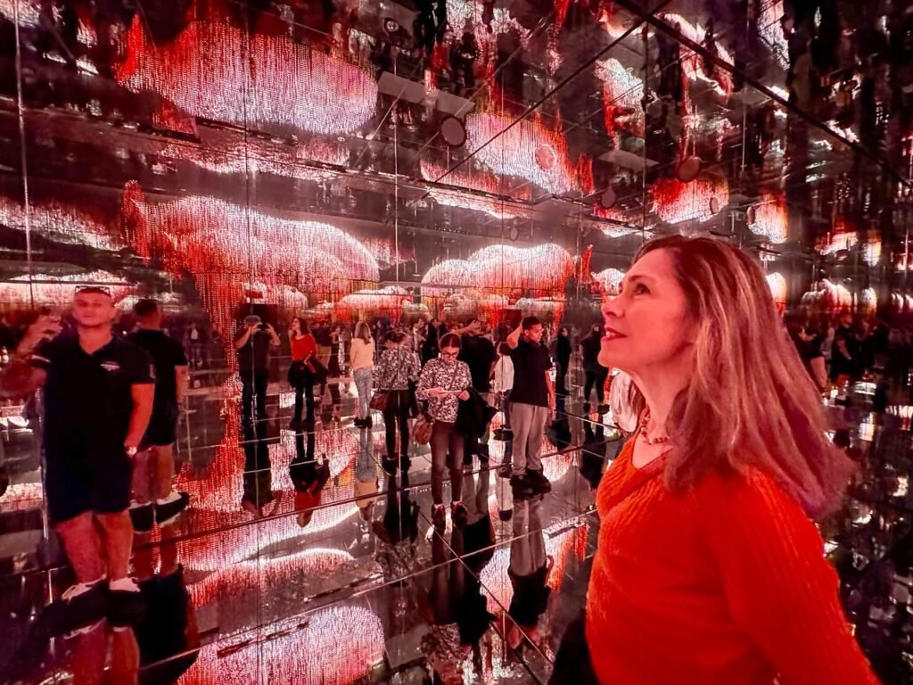 A person in a red shirt stands inside an immersive mirror maze installation featuring infinite reflections and vibrant red lighting.