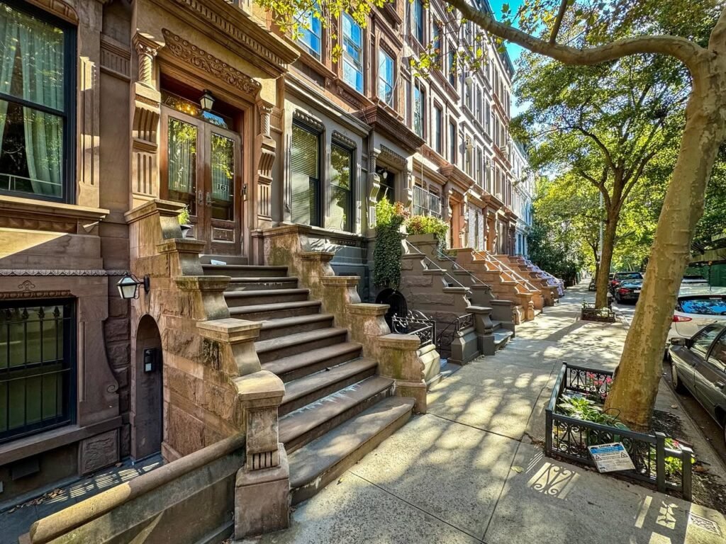 A row of brownstone buildings with grand front steps and a single street tree in a parkway.