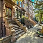 A row of brownstone buildings with grand front steps and a single street tree in a parkway.