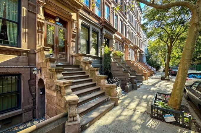 A row of brownstone buildings with grand front steps and a single street tree in a parkway.
