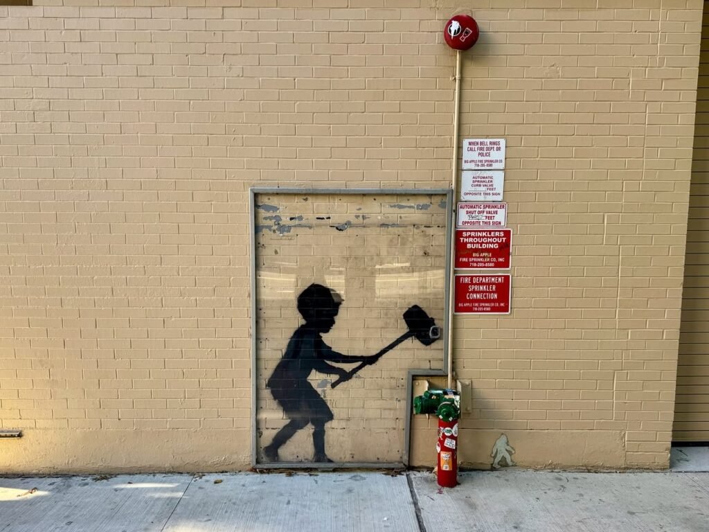 Black and white street art silhouette of a figure swinging a mallet on a beige brick wall near a fire hydrant.