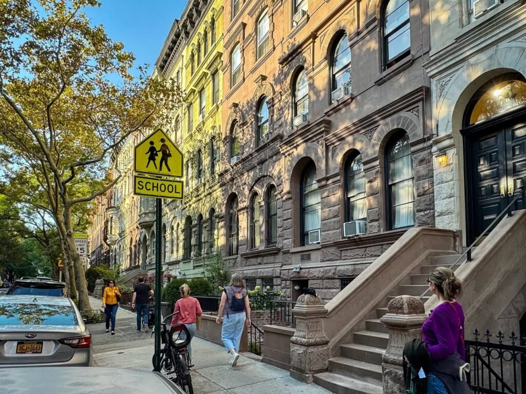 A charming street scene featuring historic brownstone buildings, pedestrians, and a yellow school crossing sign against a tree-lined background.