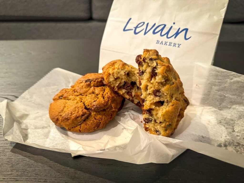 A chocolate chip cookie and a round cookie are displayed on parchment paper in a Levain Bakery bag. The treats rest on a wooden surface in a casual setting.