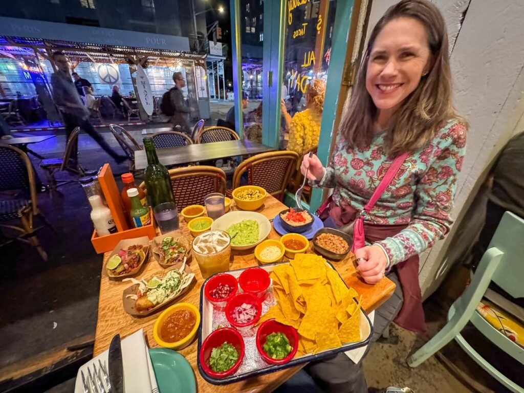 A smiling person at a taco shop table enjoying nachos, guacamole, and street tacos while staff work in the background.