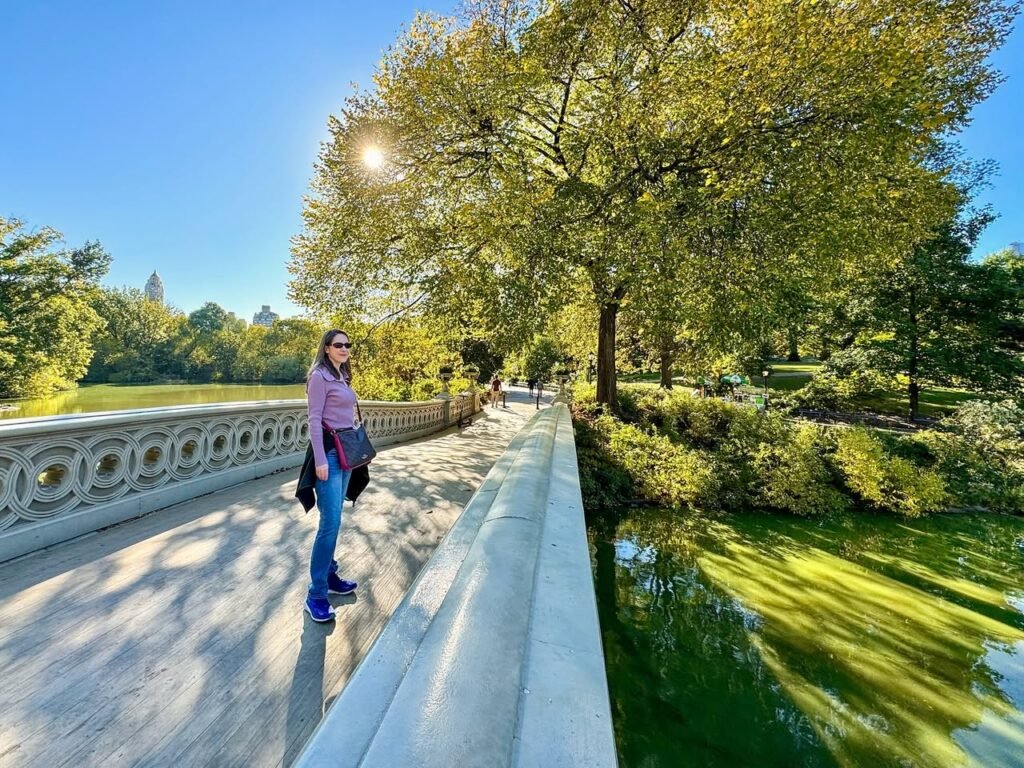A visitor stands on the ornate balustrade of a stone bridge beside a green reflecting pond under a clear blue sky.
