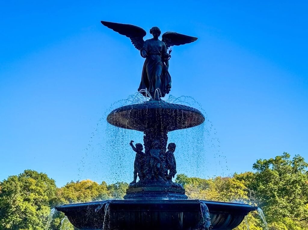 A grand bronze fountain topped with an angel statue, featuring multiple tiers and cascading water under a clear blue sky.