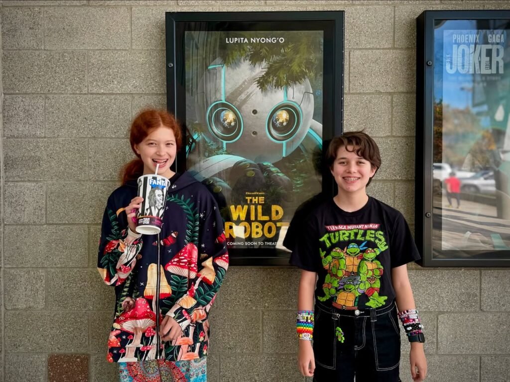 Two people stand smiling in front of a cinema wall featuring posters for The Wild Robot and The Joker.