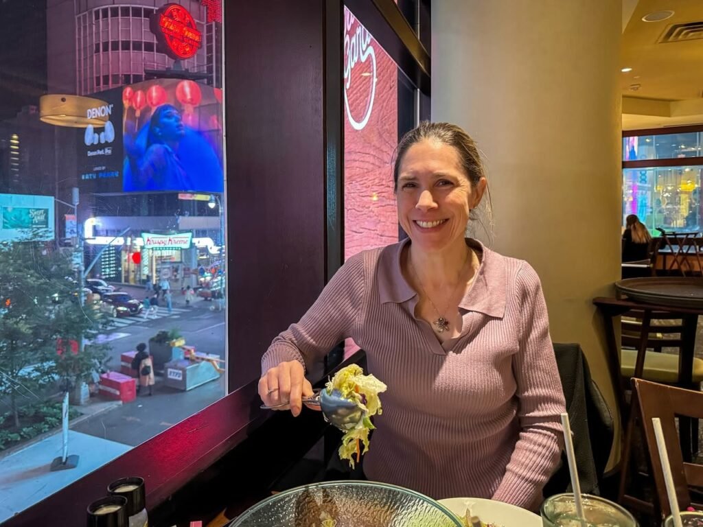 A woman enjoying a meal at a restaurant with a view of the city lights through a large window. The background features a bright urban scene with digital displays.