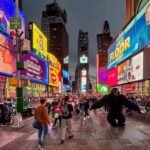 A bustling street scene in Times Square, New York City, featuring bright digital billboards and costumed performers.