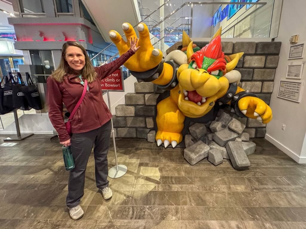 A person poses for a photo with a large Bowser statue displayed in a retail store. The setting features the character surrounded by stone props, capturing a playful moment.